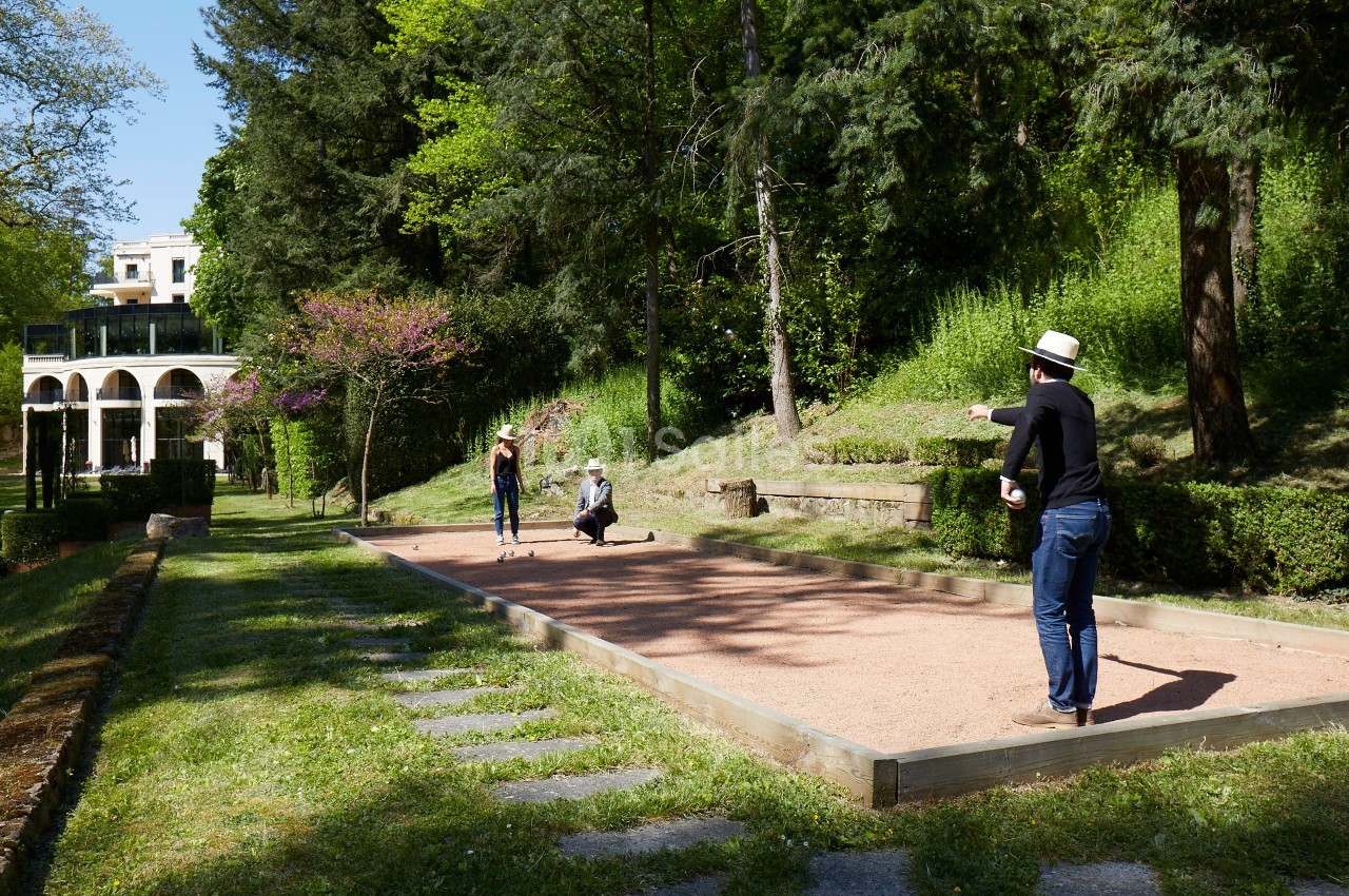 Personnes jouant à la pétanque sur un terrain ensoleillé entouré de verdure et d'arbres.