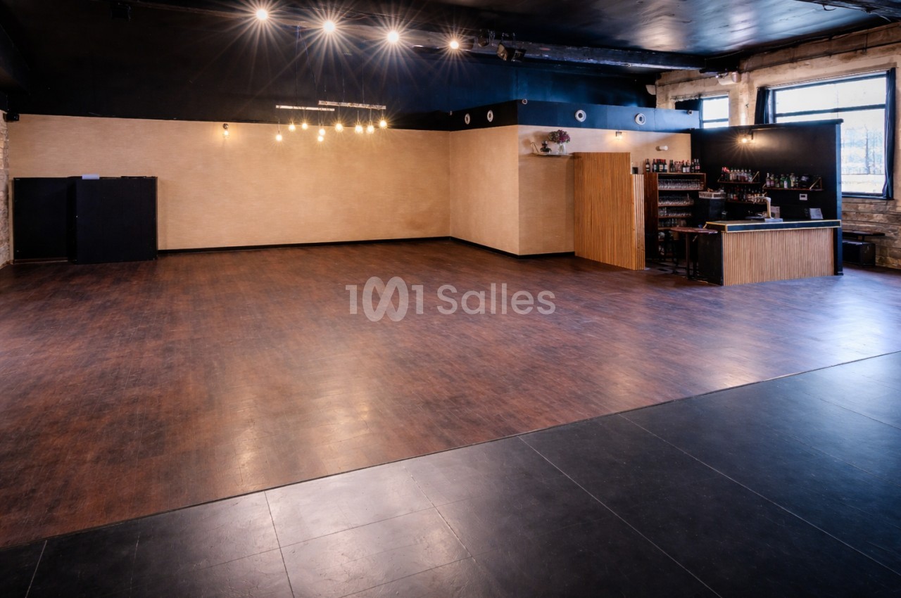 Salle vide avec parquet, éclairage suspendu, bar en bois à droite et murs clairs dans un espace industriel.