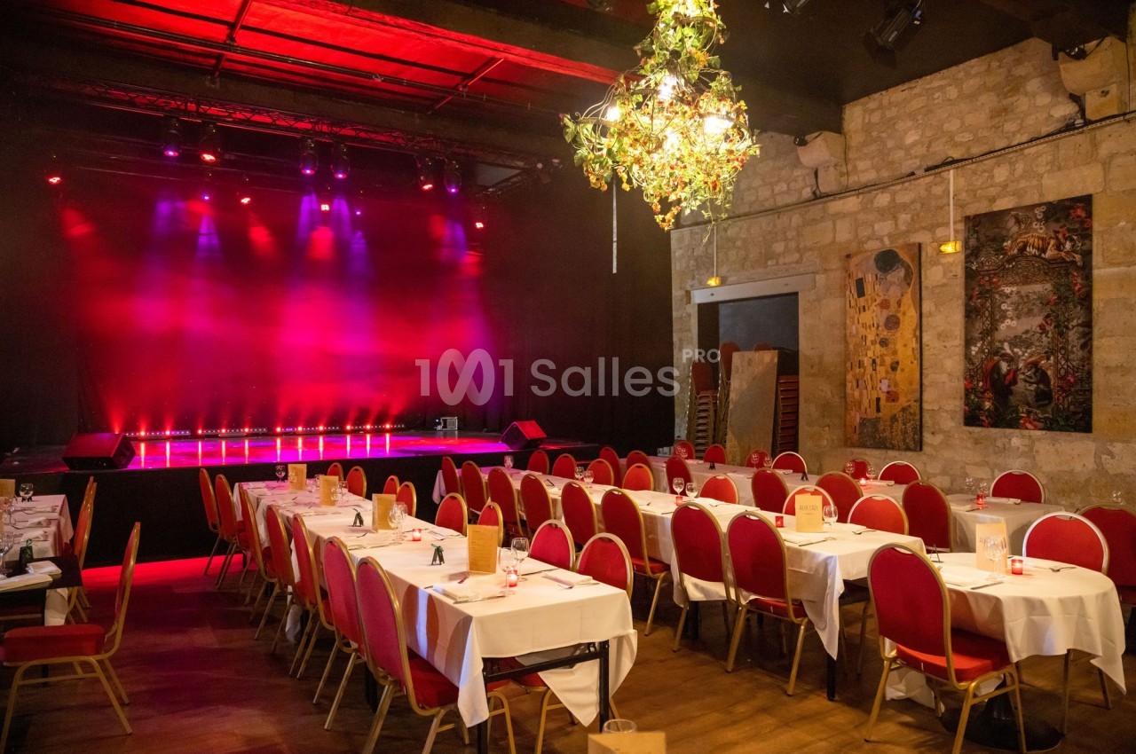 Salle de spectacle avec tables dressées pour un dîner, éclairage rouge sur la scène et décoration suspendue.