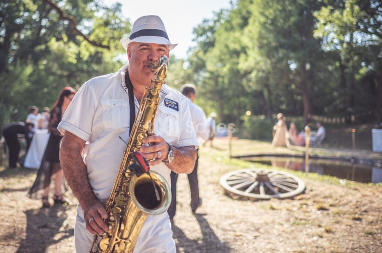 Un homme en tenue blanche joue du saxophone en extérieur, près d'un étang et sous un ciel ensoleillé.