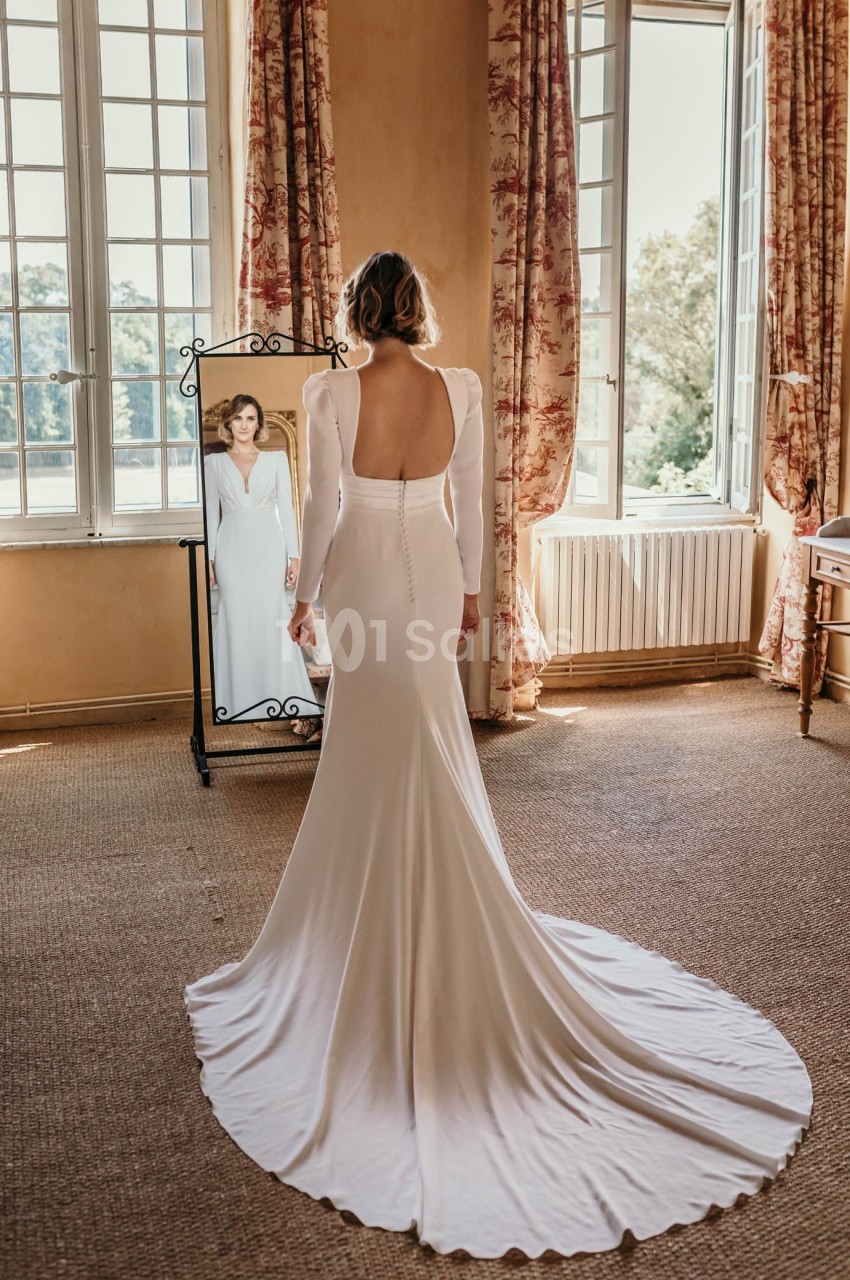 Une femme en robe de mariée blanche se regarde dans un miroir dans une pièce lumineuse avec de grandes fenêtres.