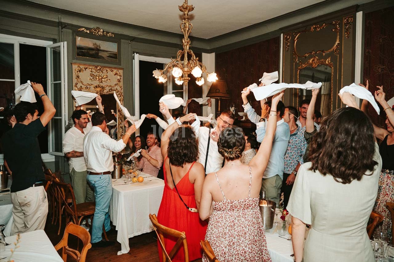 Des convives célèbrent autour d'une table dans une salle élégante, levant des serviettes en l'air dans une ambiance festive.