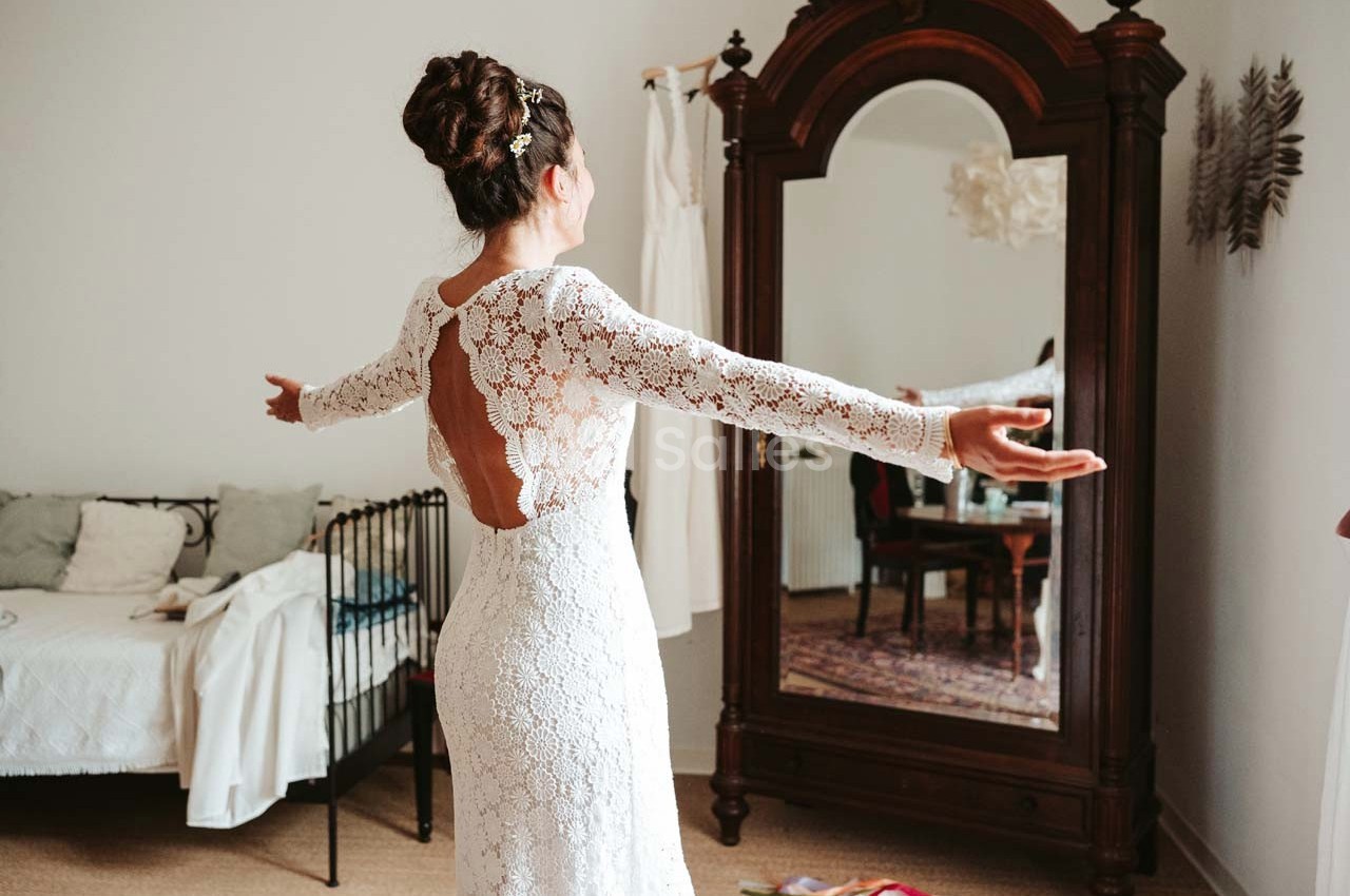 Femme en robe de mariée en dentelle blanche, bras ouverts, devant un miroir ancien dans une chambre lumineuse.