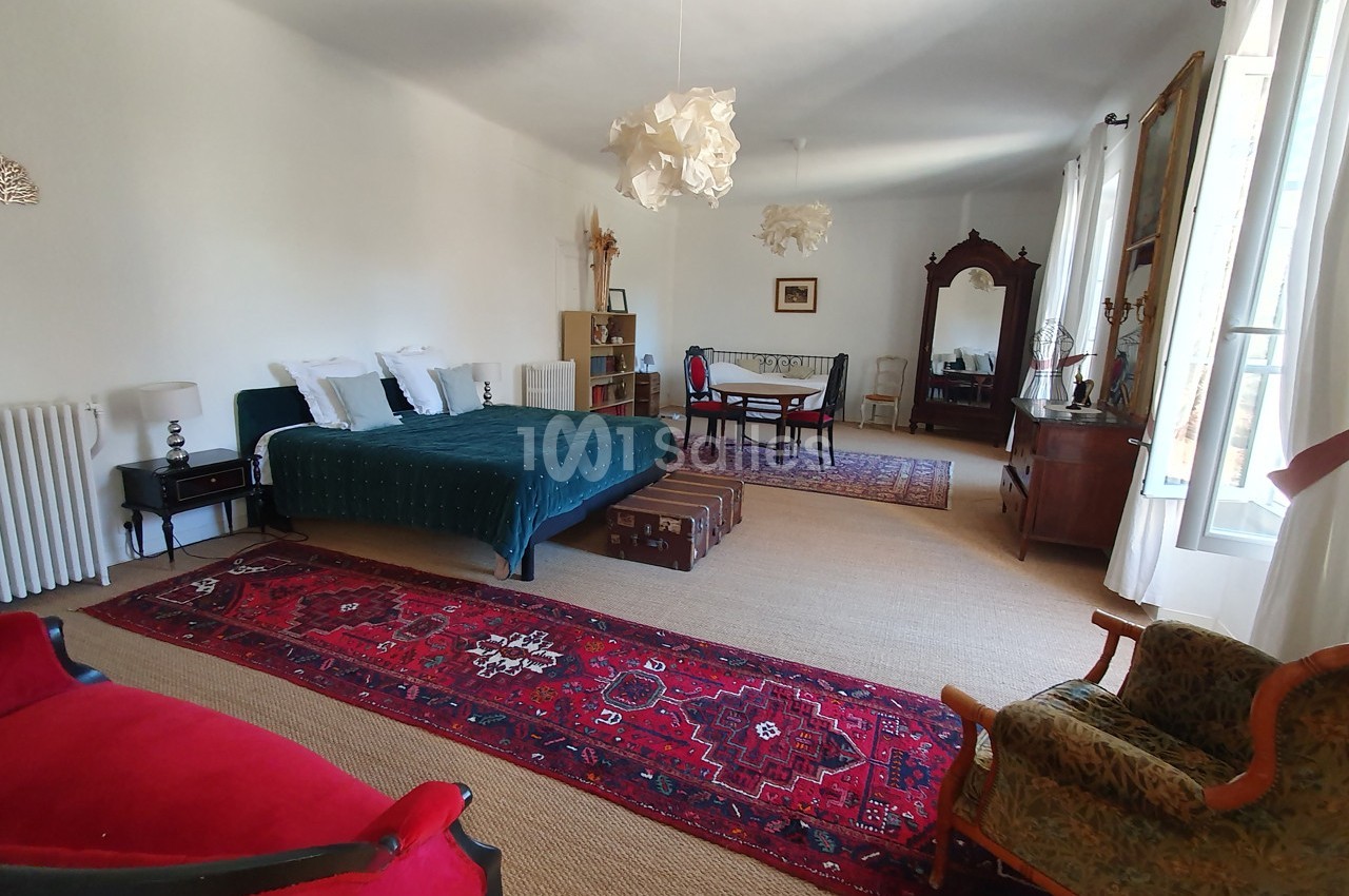 Grande chambre lumineuse avec lit double, tapis rouges, meubles anciens, coin repas et grandes fenêtres.