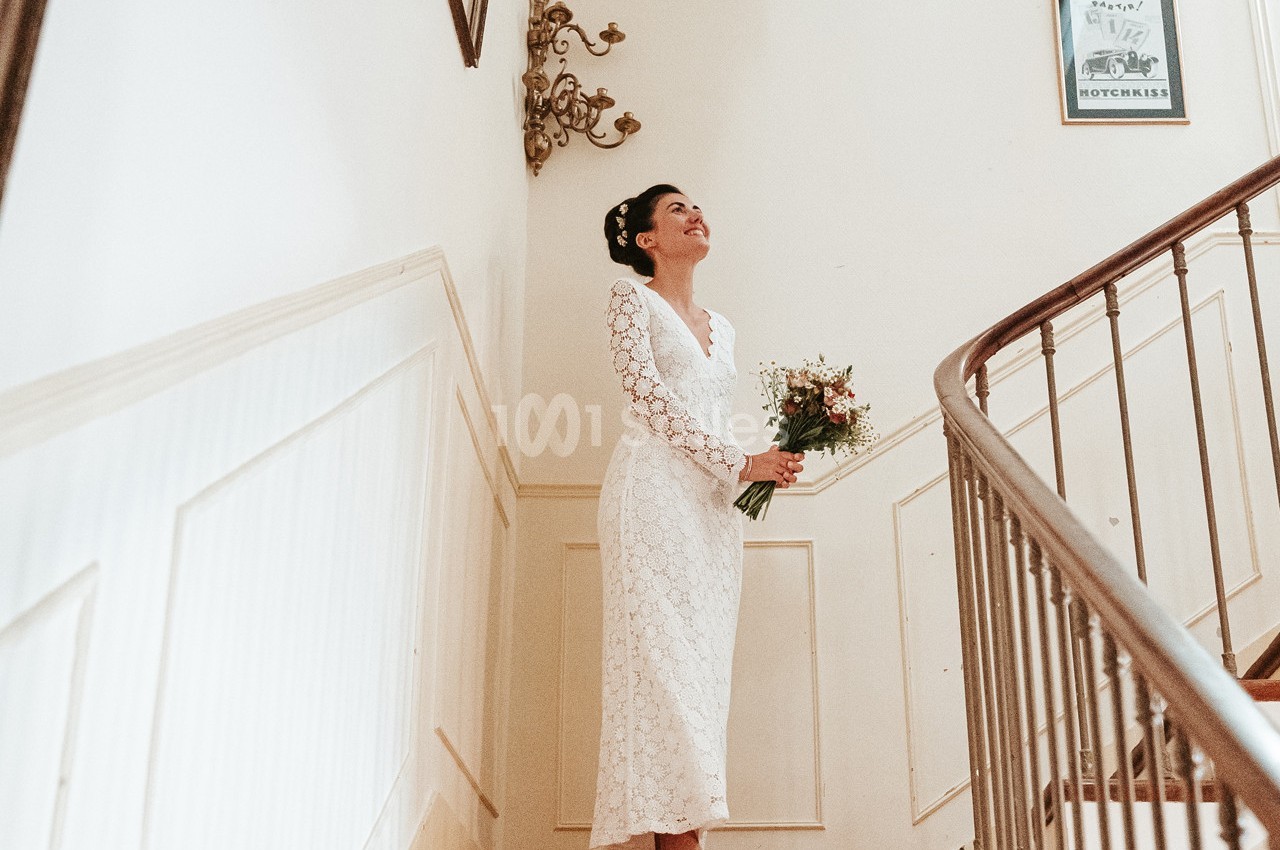 Une femme en robe blanche tient un bouquet de fleurs en haut d'un escalier dans un intérieur lumineux.