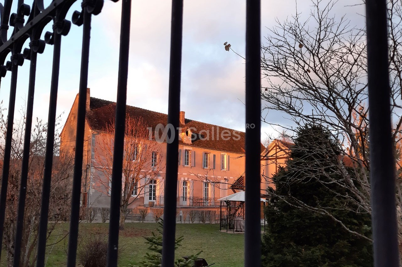 Façade d'une maison en pierre rose vue à travers une grille, entourée d'arbres et d'un jardin au crépuscule.