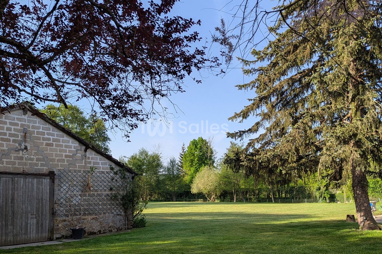 Vue d'un jardin verdoyant avec pelouse, arbres et une petite dépendance en pierre sous un ciel dégagé.