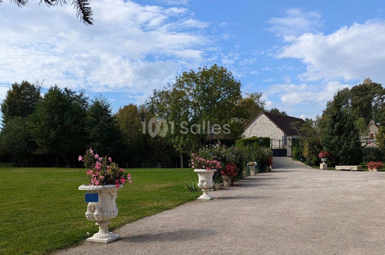 Allée bordée de jardinières fleuries menant à une maison en pierre entourée de verdure sous un ciel partiellement nuageux.