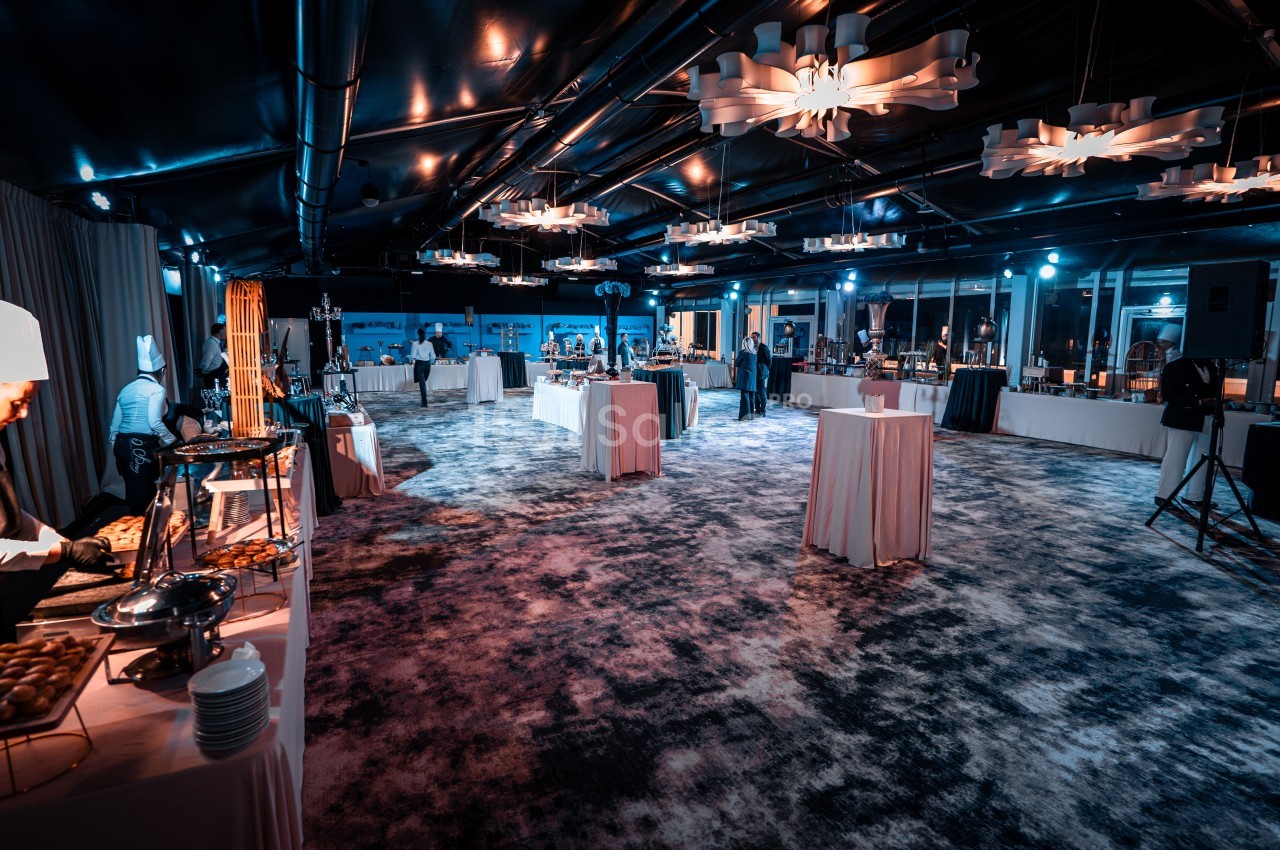 Salle de réception éclairée avec des buffets dressés, des tables hautes et des chefs en train de préparer des plats.