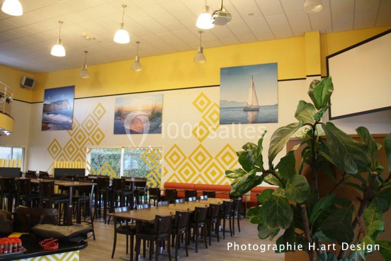 Salle lumineuse avec tables et chaises en bois, murs décorés de motifs jaunes et tableaux représentant des paysages.