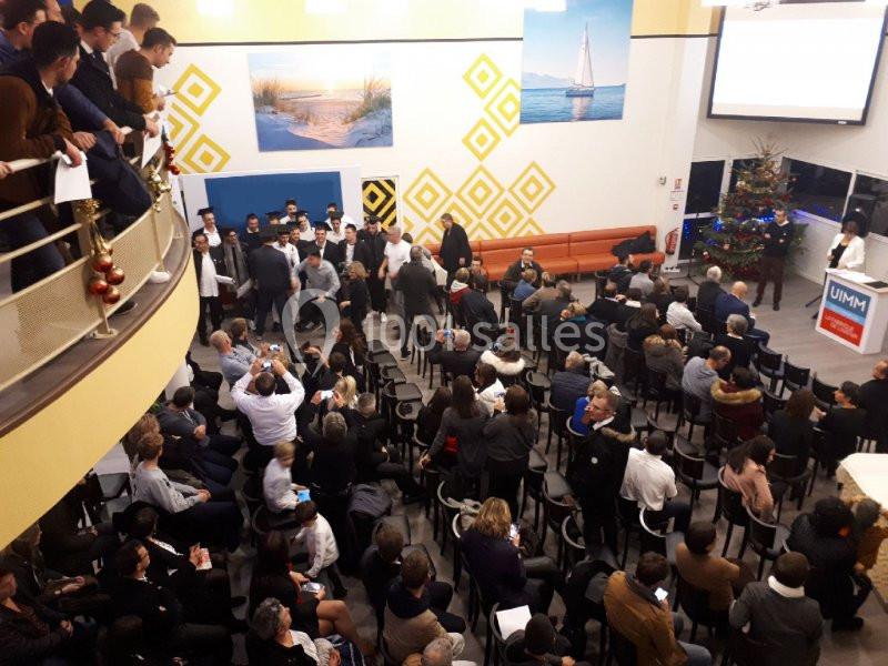 Salle comble avec des participants assis et debout lors d'un événement, décorée avec un sapin de Noël et des tableaux muraux.