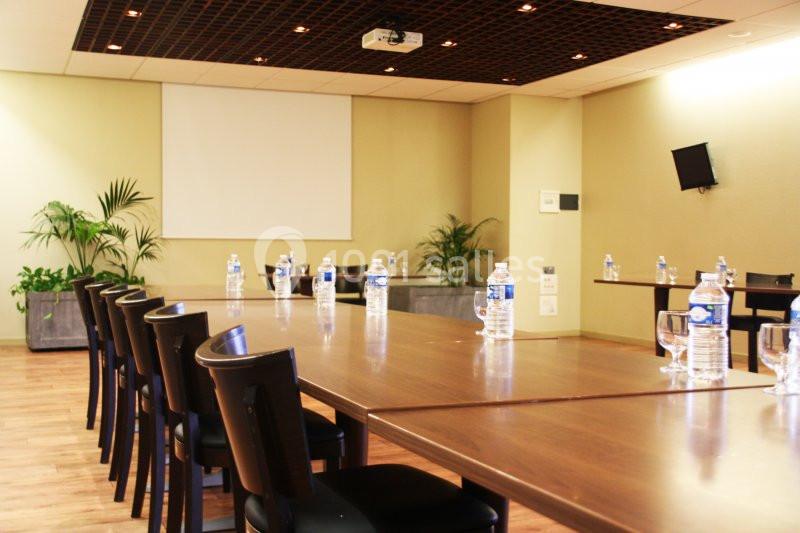 Salle de réunion lumineuse avec tables en bois, chaises alignées, bouteilles d'eau et écran de projection.