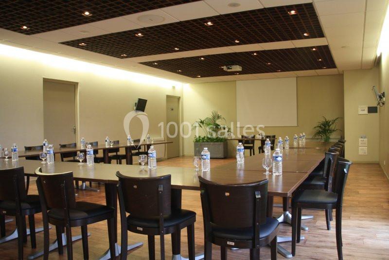Salle de réunion avec tables disposées en U, chaises, bouteilles d'eau, écran de projection et plantes en arrière-plan.
