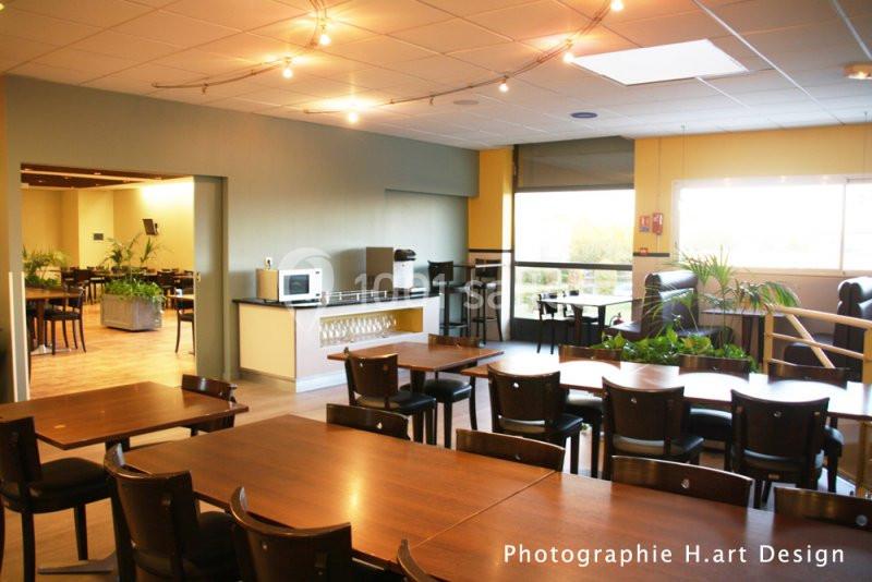 Salle lumineuse avec tables en bois, chaises noires, coin cuisine équipé et grandes fenêtres donnant sur l'extérieur.