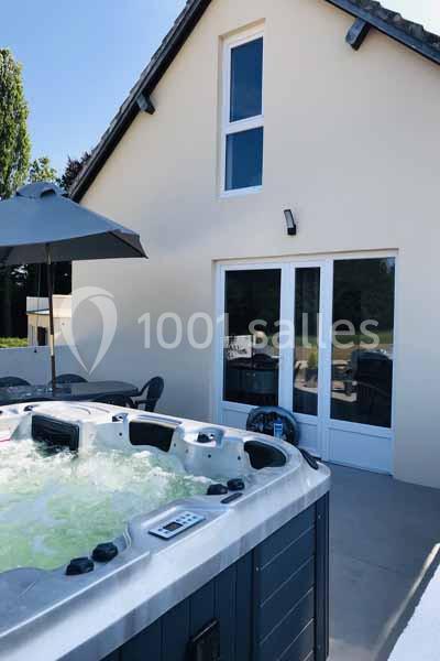 Spa bouillonnant sur une terrasse ensoleillée devant une maison avec baies vitrées et parasol.