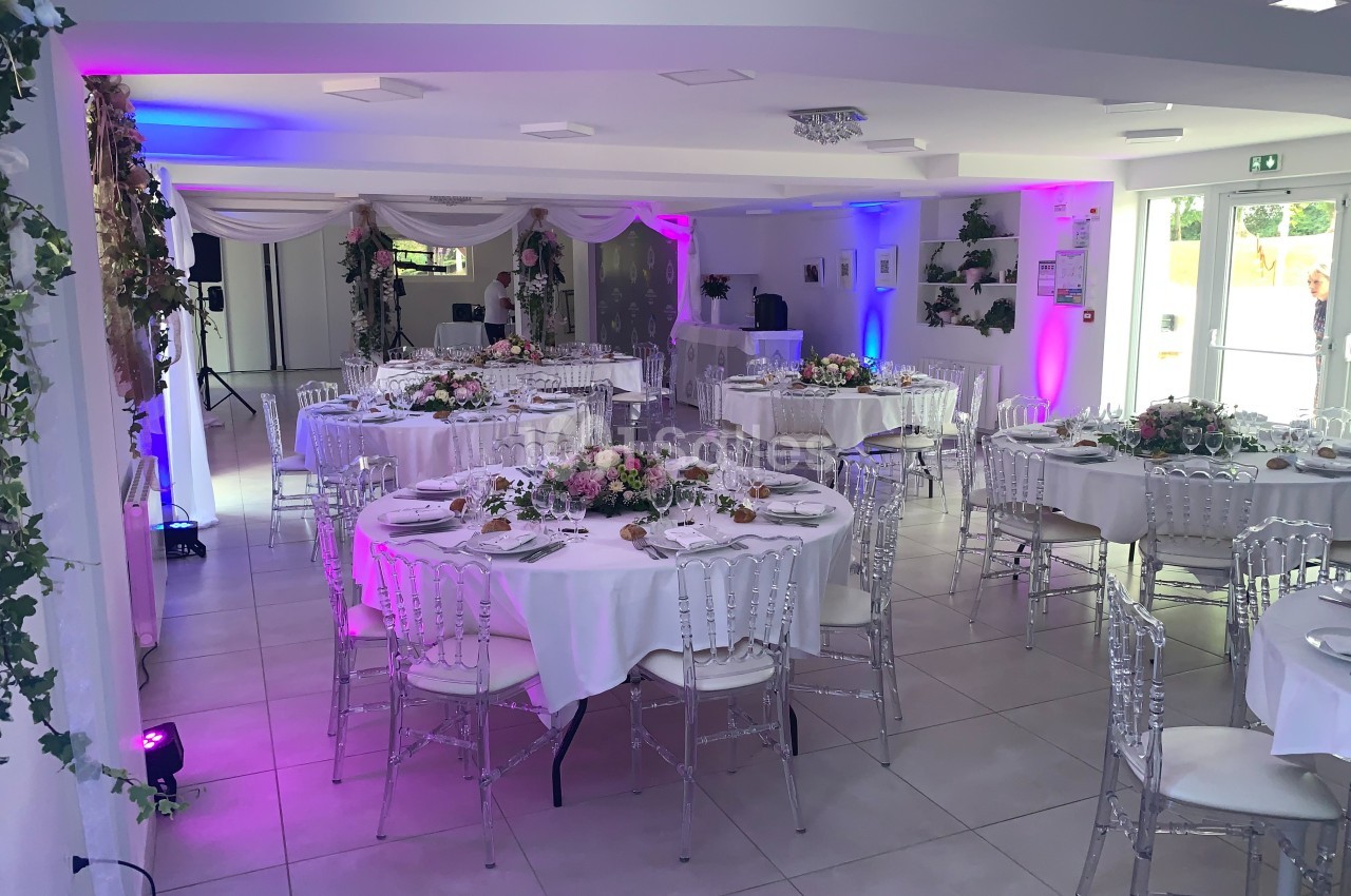Salle de réception décorée avec des tables rondes dressées, des chaises transparentes et des éclairages colorés.