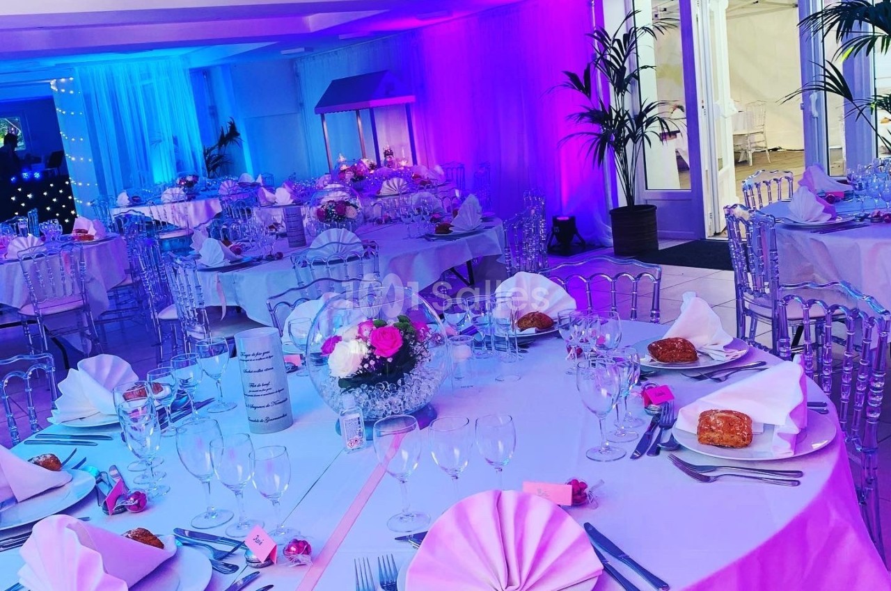 Salle de réception décorée avec des tables rondes, nappes blanches, fleurs et éclairage aux tons rose et bleu.