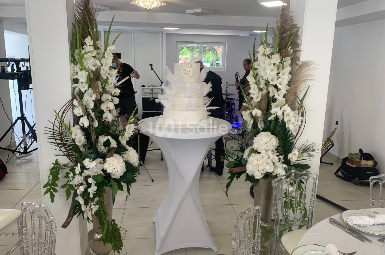 Gâteau de mariage blanc à plusieurs étages sur une table, entouré de grandes compositions florales blanches.