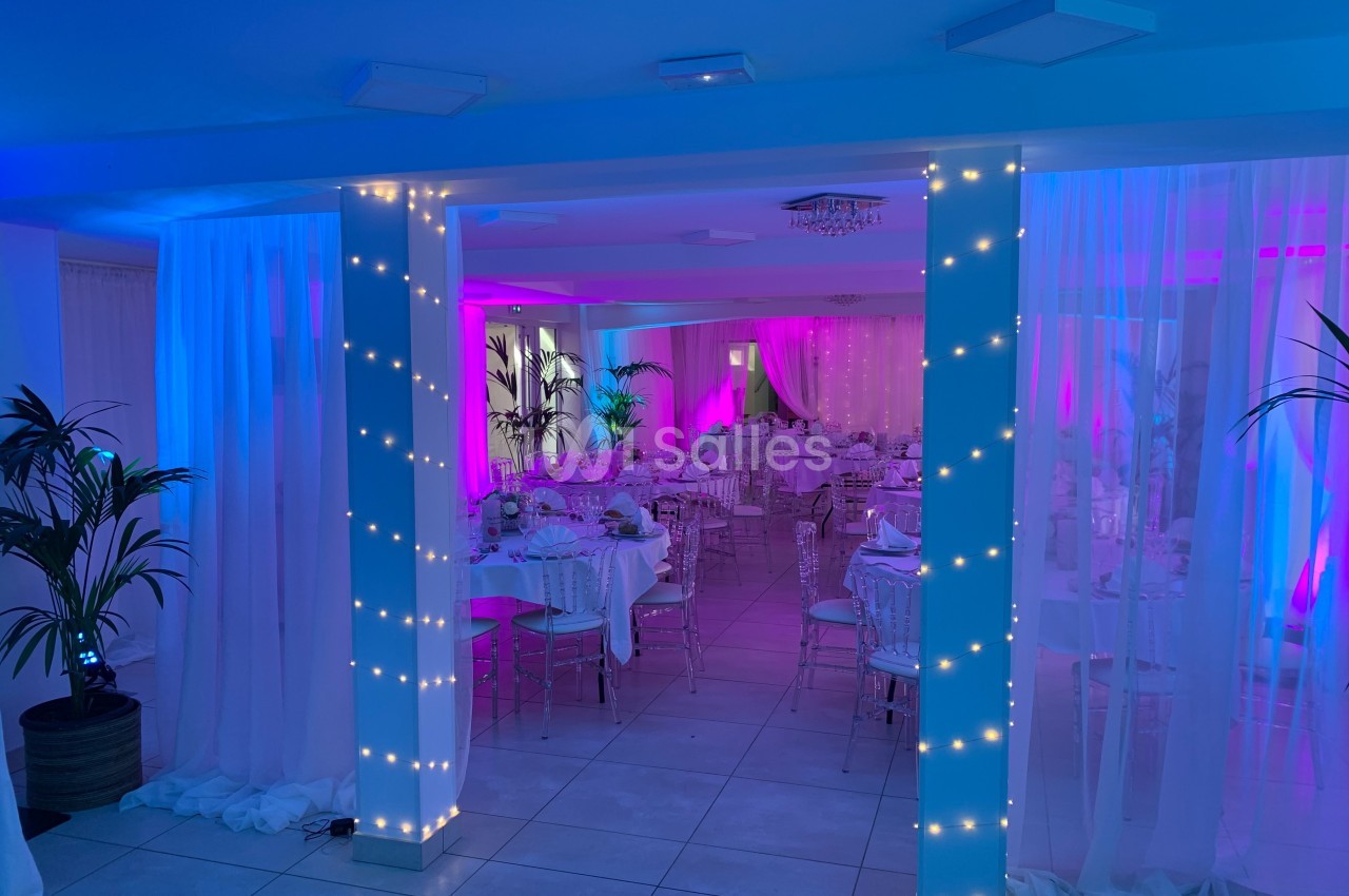 Salle de réception décorée avec des guirlandes lumineuses, des tables dressées et un éclairage rose et bleu.