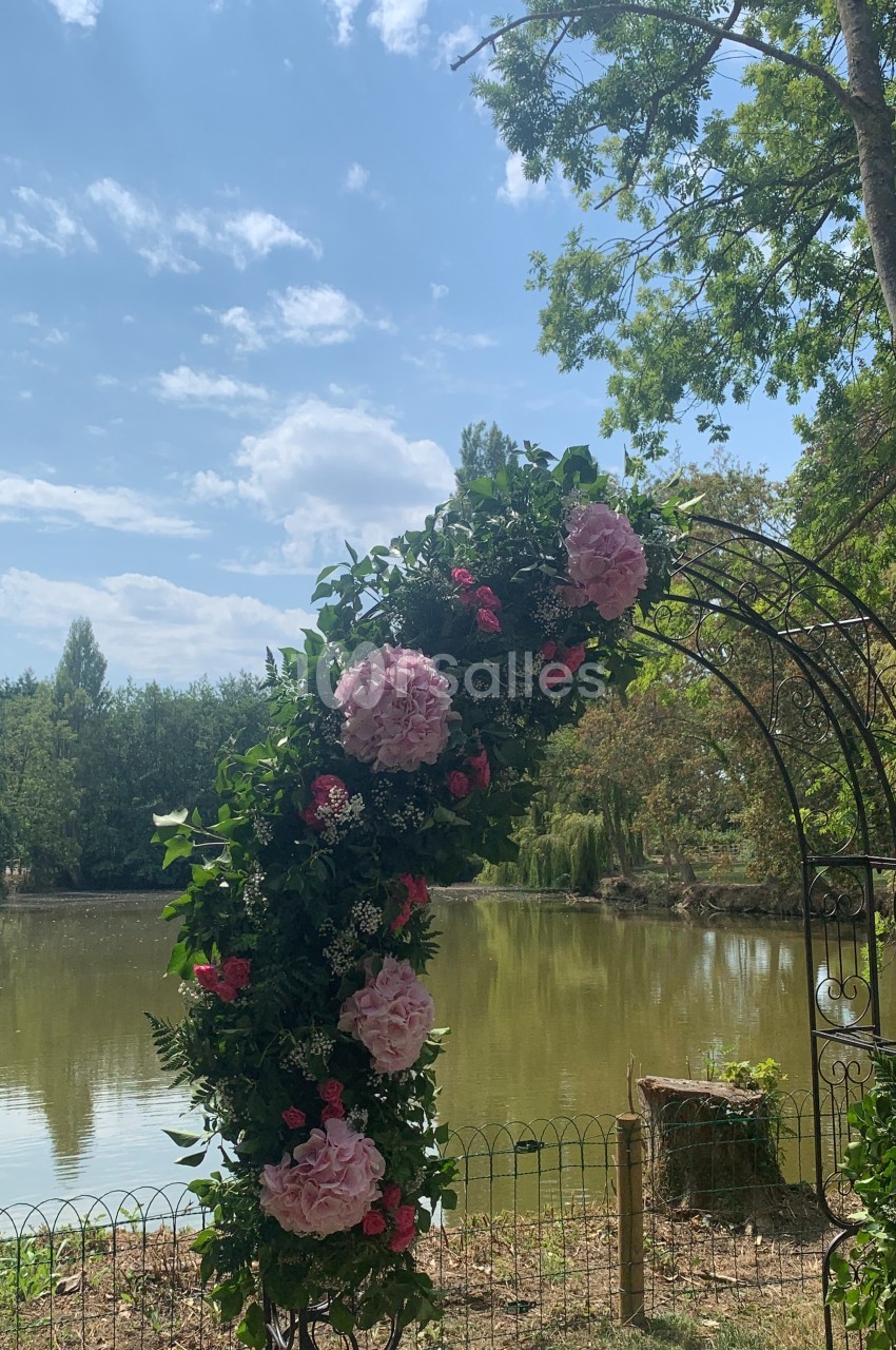 Arche décorée de fleurs roses et feuillage, placée devant un étang entouré d'arbres sous un ciel ensoleillé.