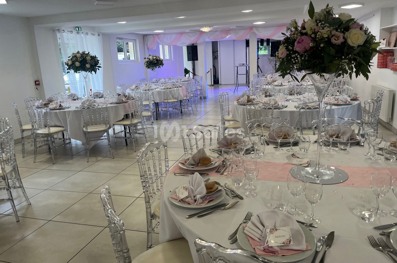 Salle de réception décorée avec des tables rondes, nappes blanches, chaises transparentes et compositions florales.