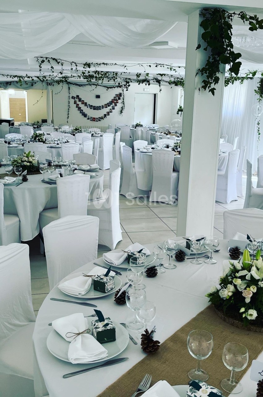 Salle de réception décorée avec des tables rondes, nappes blanches, centres de table floraux et guirlandes végétales.