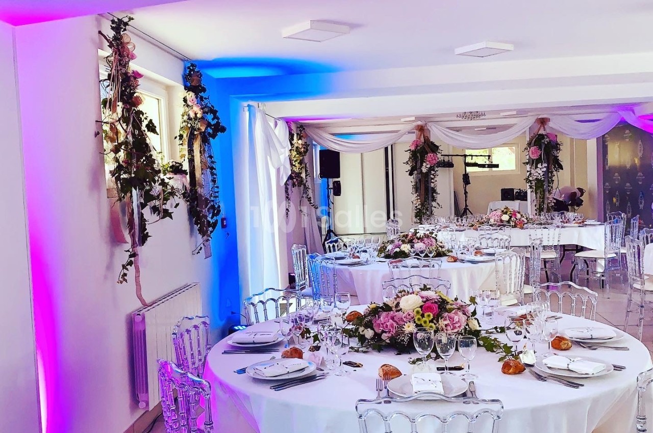 Salle de réception décorée avec des tables rondes, nappes blanches, chaises transparentes et arrangements floraux élégants.