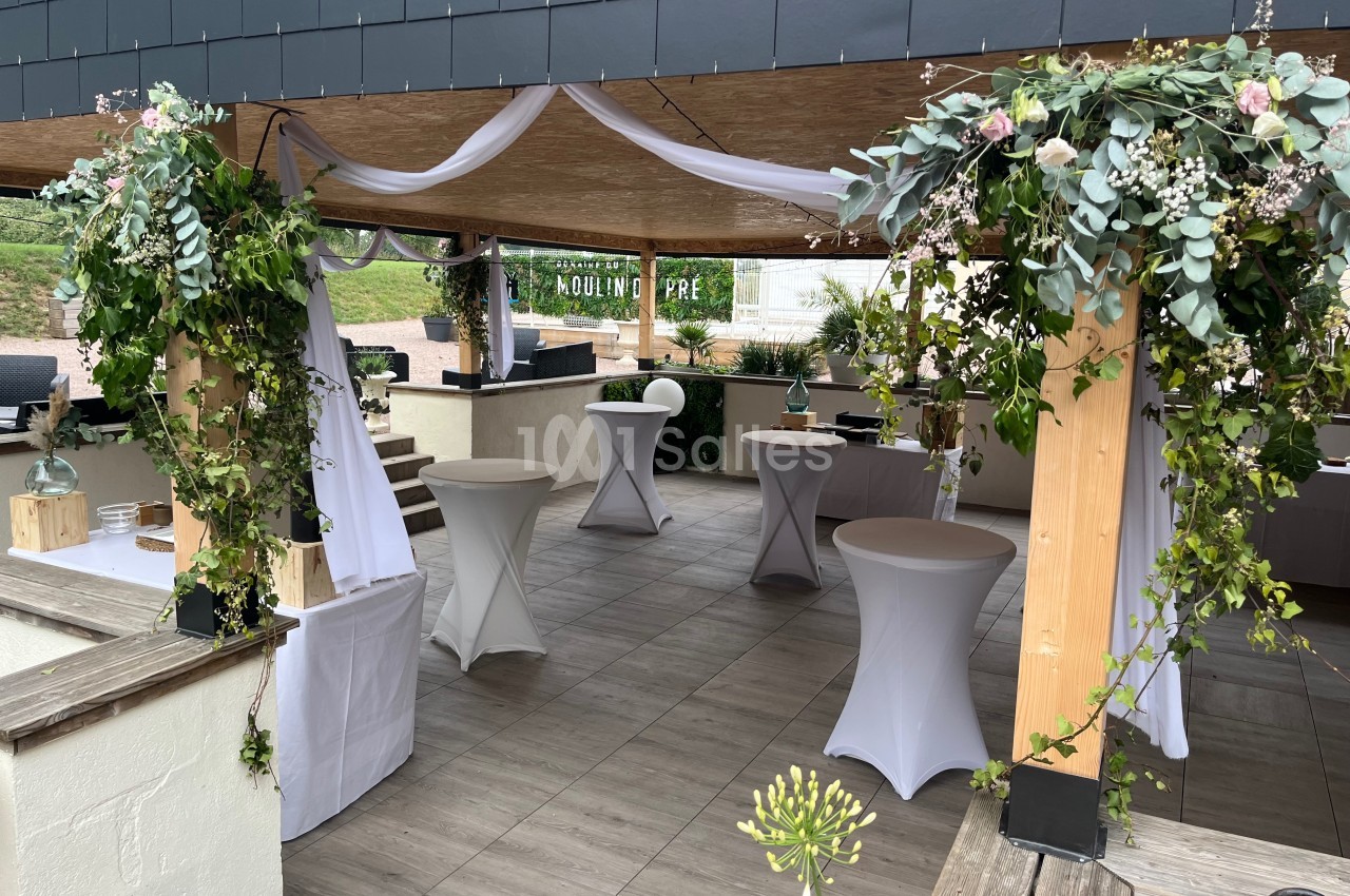 Espace extérieur décoré pour un événement, avec tables hautes, voilages blancs et arrangements floraux suspendus.