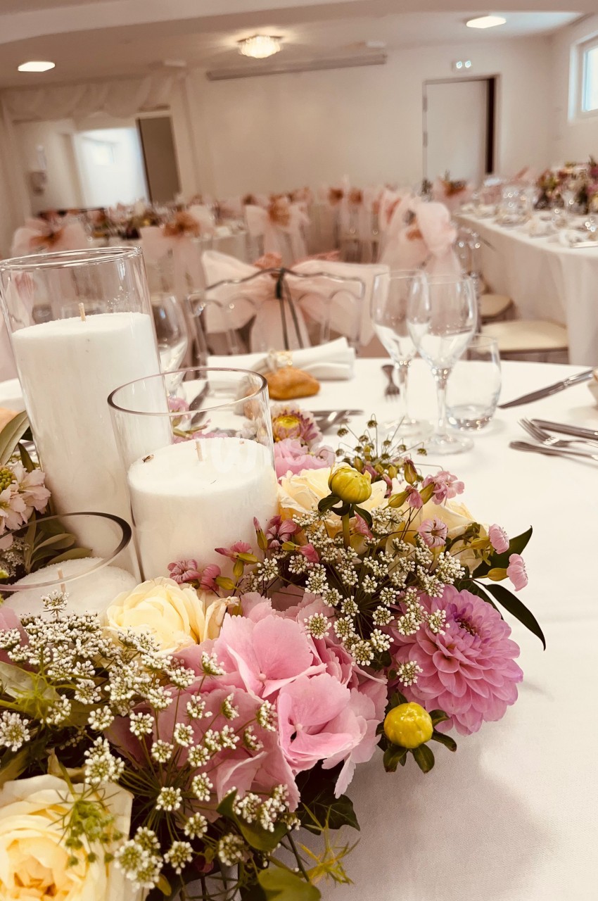 Centre de table floral avec bougies blanches, entouré de vaisselle et nappes blanches dans une salle de réception.
