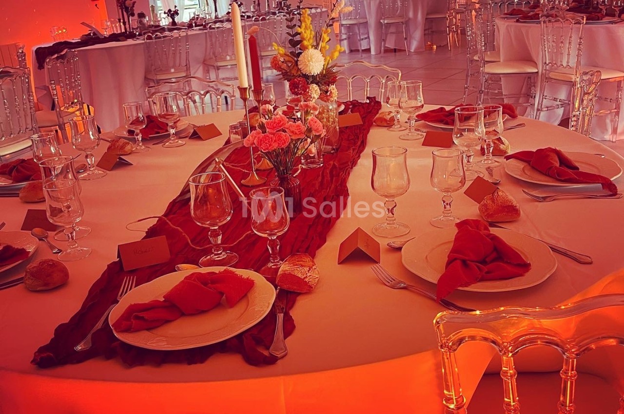 Table ronde décorée pour un événement, avec nappes blanches, serviettes rouges, chandeliers et fleurs au centre.