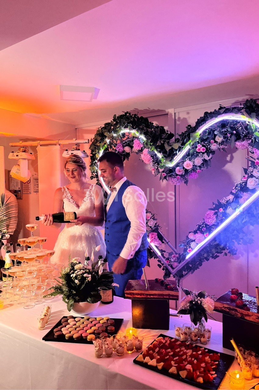 Un couple de mariés coupe un gâteau devant une décoration lumineuse en forme de cœur, entouré de desserts.
