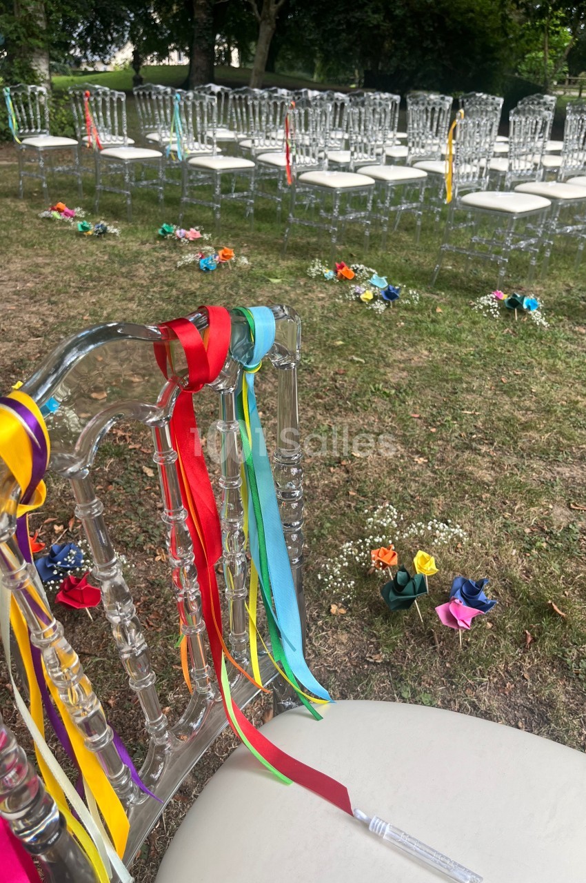 Chaises transparentes décorées de rubans colorés alignées en extérieur, entourées de fleurs disposées sur l'herbe.