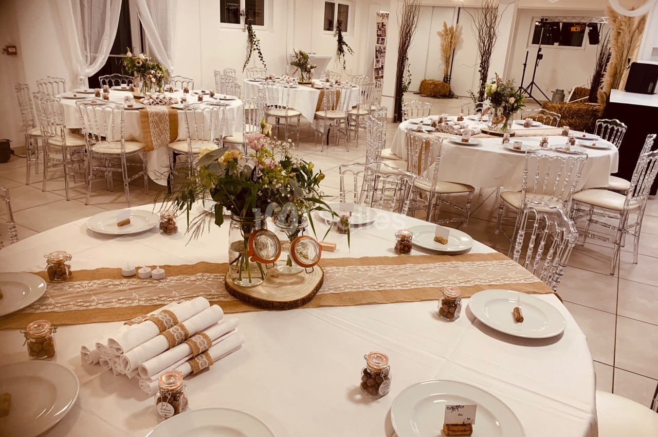 Salle décorée pour un événement, avec tables rondes dressées, nappes blanches, décorations florales et éléments en toile de…
