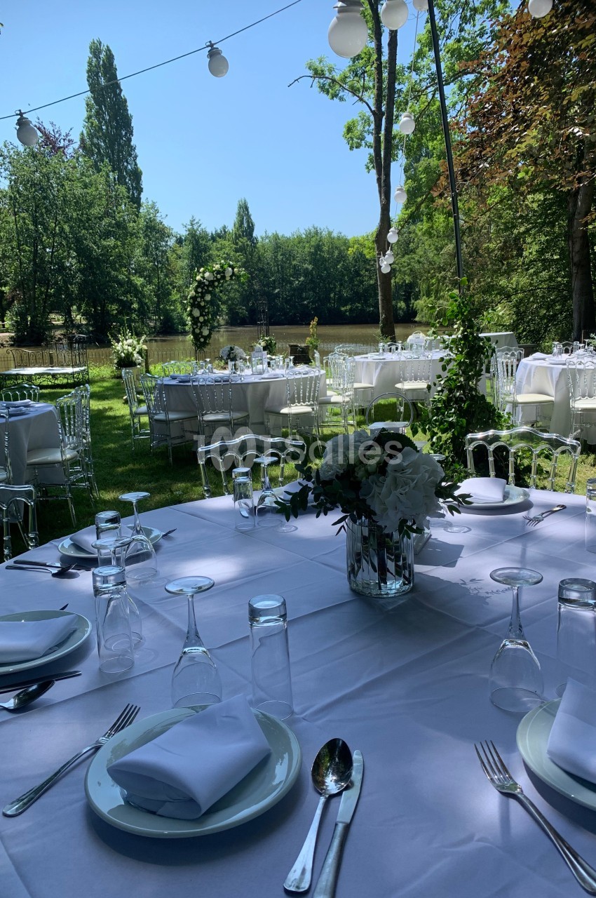 Tables élégamment dressées en extérieur, entourées de verdure et décorées de fleurs blanches sous des guirlandes lumineuses.