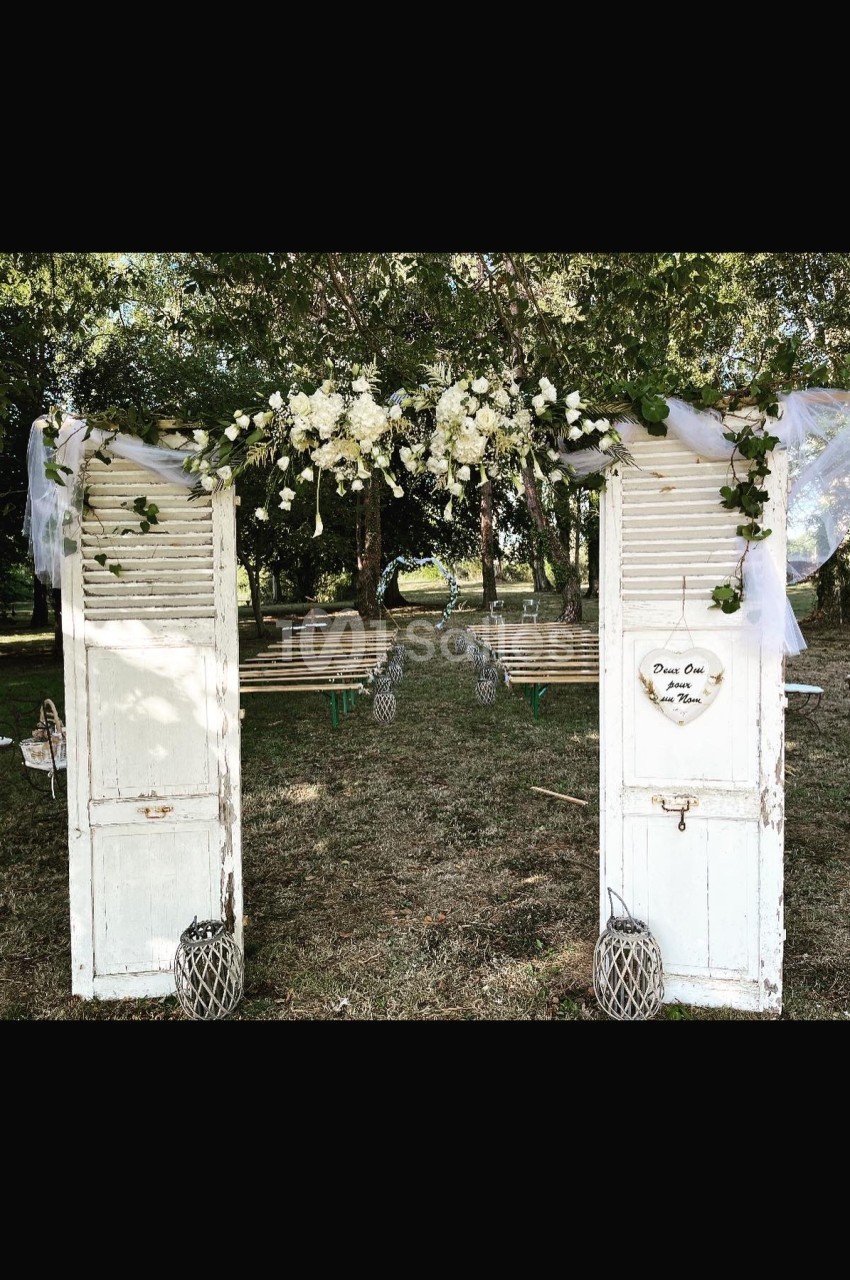 Deux portes blanches anciennes décorées de fleurs blanches et de voilages, encadrant une allée extérieure.
