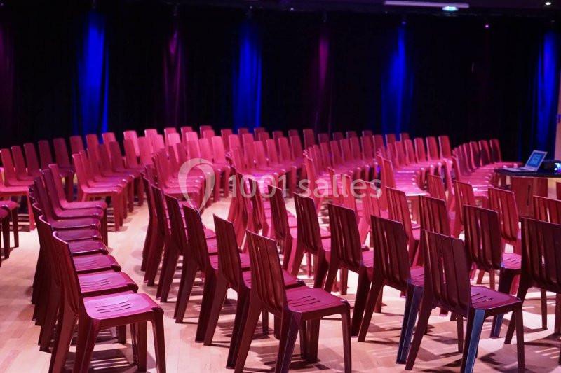 Salle avec des rangées de chaises rouges en plastique alignées sur un sol en bois, éclairage coloré en arrière-plan.