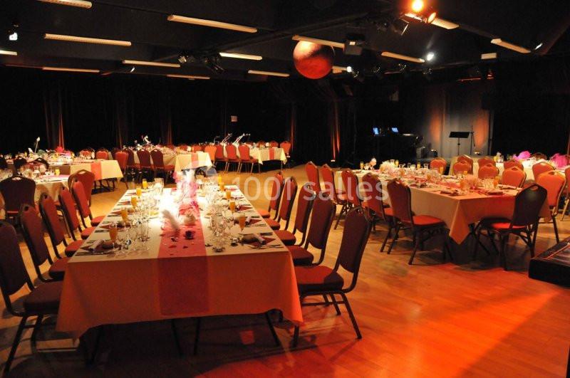 Salle de réception éclairée avec des tables dressées pour un événement, entourées de chaises rouges.