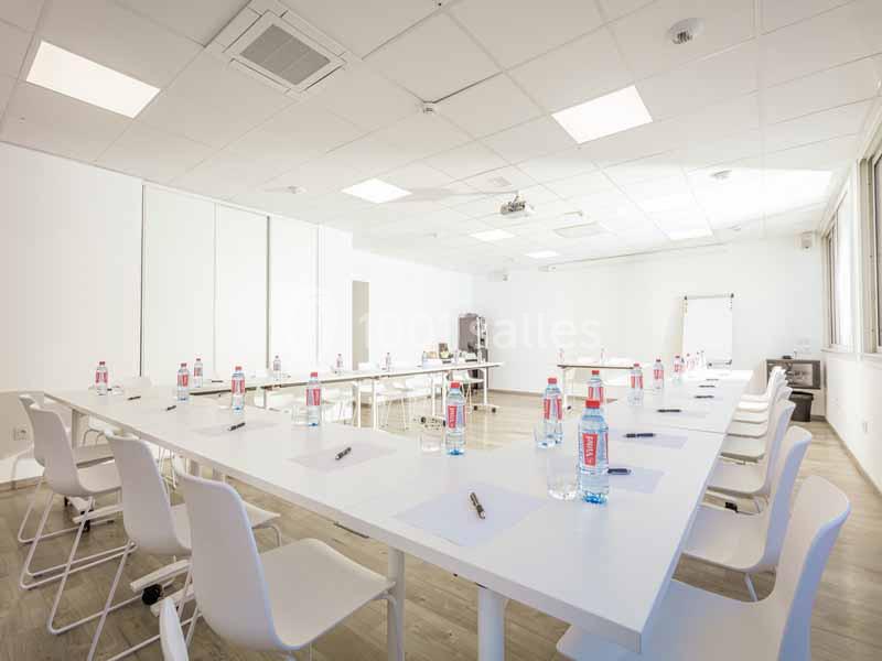 Salle de réunion lumineuse avec tables blanches disposées en U, chaises, bouteilles d'eau et matériel de prise de notes.
