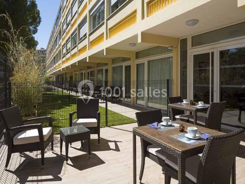 Terrasse extérieure avec tables et chaises en rotin, entourée de verdure et attenante à un bâtiment moderne.