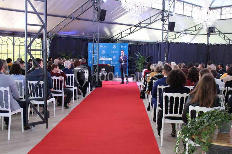 Un conférencier s'adresse à un public assis dans une salle sous chapiteau avec tapis rouge et décorations.