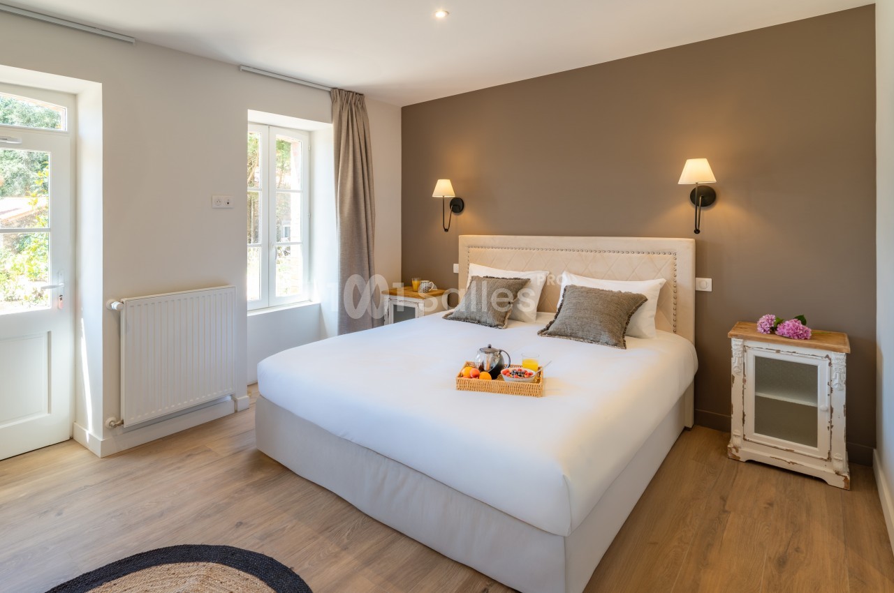 Chambre lumineuse avec lit double, murs beige et taupe, tables de chevet, et plateau de fruits sur le lit.