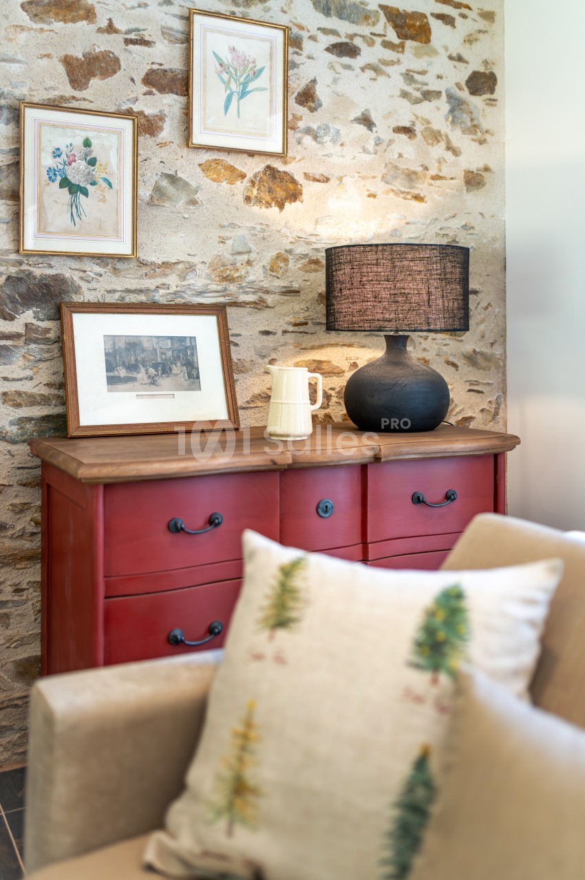 Commode rouge surmontée de cadres, lampe noire et carafe, avec un mur en pierre en arrière-plan.