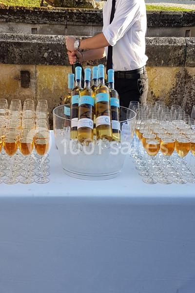 Bouteilles de vin blanc sur glace entourées de verres remplis d'un liquide doré, disposées sur une table blanche.
