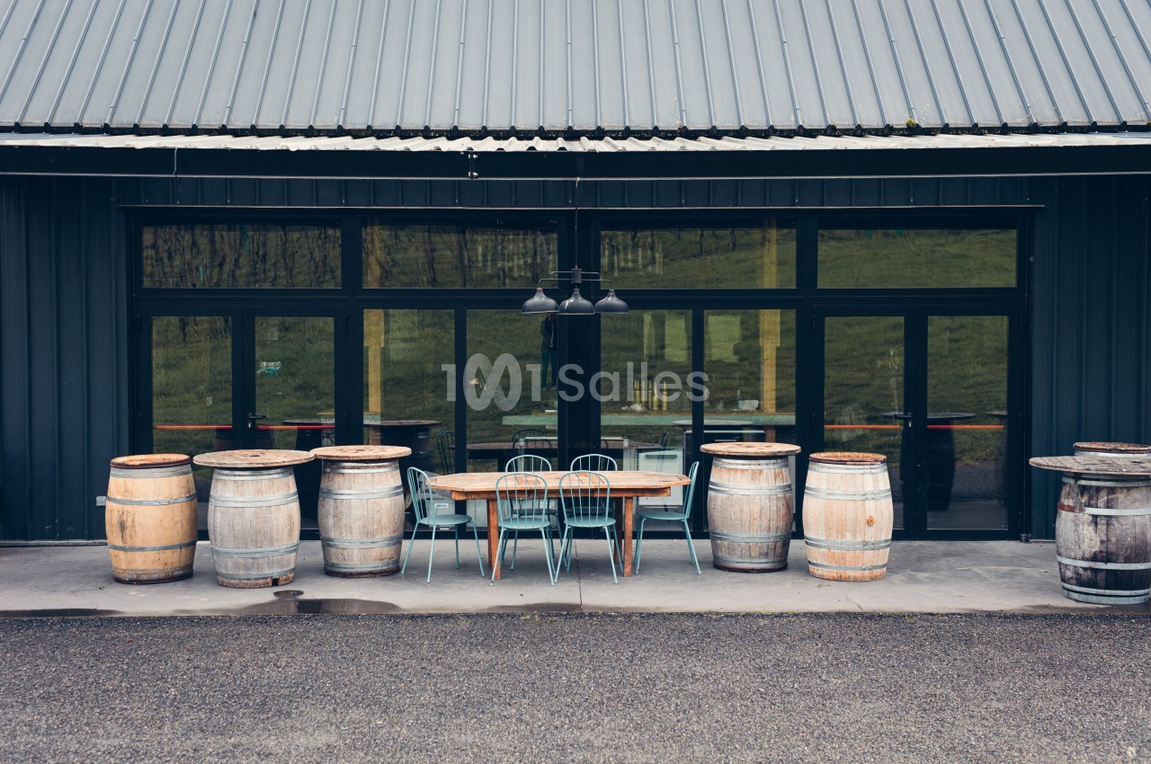 Façade d'un bâtiment avec une table en bois, des chaises métalliques et des tonneaux disposés sur une terrasse en béton.