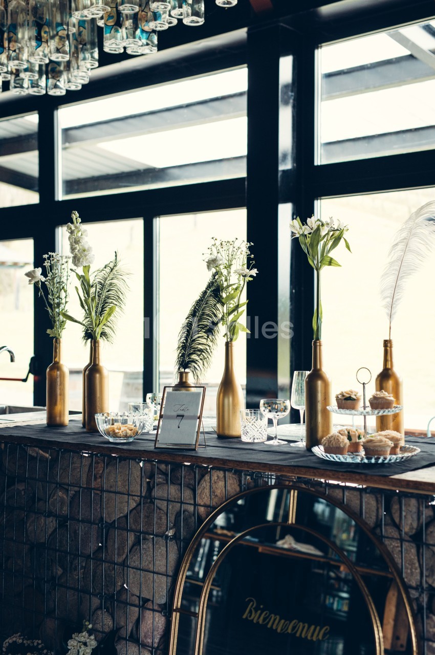 Décoration de table avec bouteilles dorées contenant des fleurs, plumes et vaisselle dans un cadre lumineux.