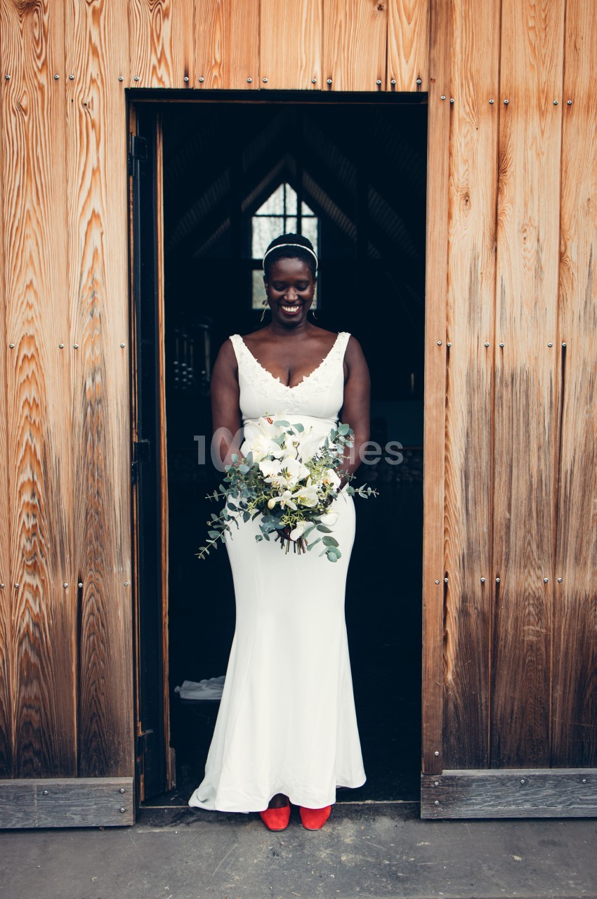 Femme en robe de mariée blanche tenant un bouquet de fleurs, debout dans l'encadrement d'une porte en bois.