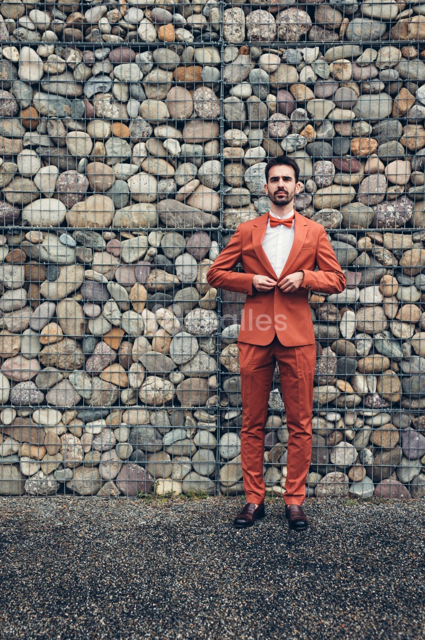 Homme en costume orange ajustant sa veste devant un mur de pierres grillagées.