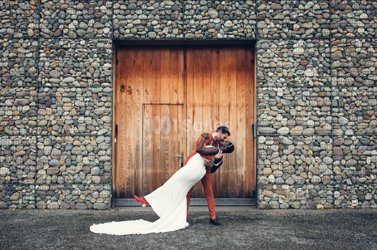 Un couple en tenue de mariage s'embrasse devant une porte en bois encadrée par un mur de pierres.