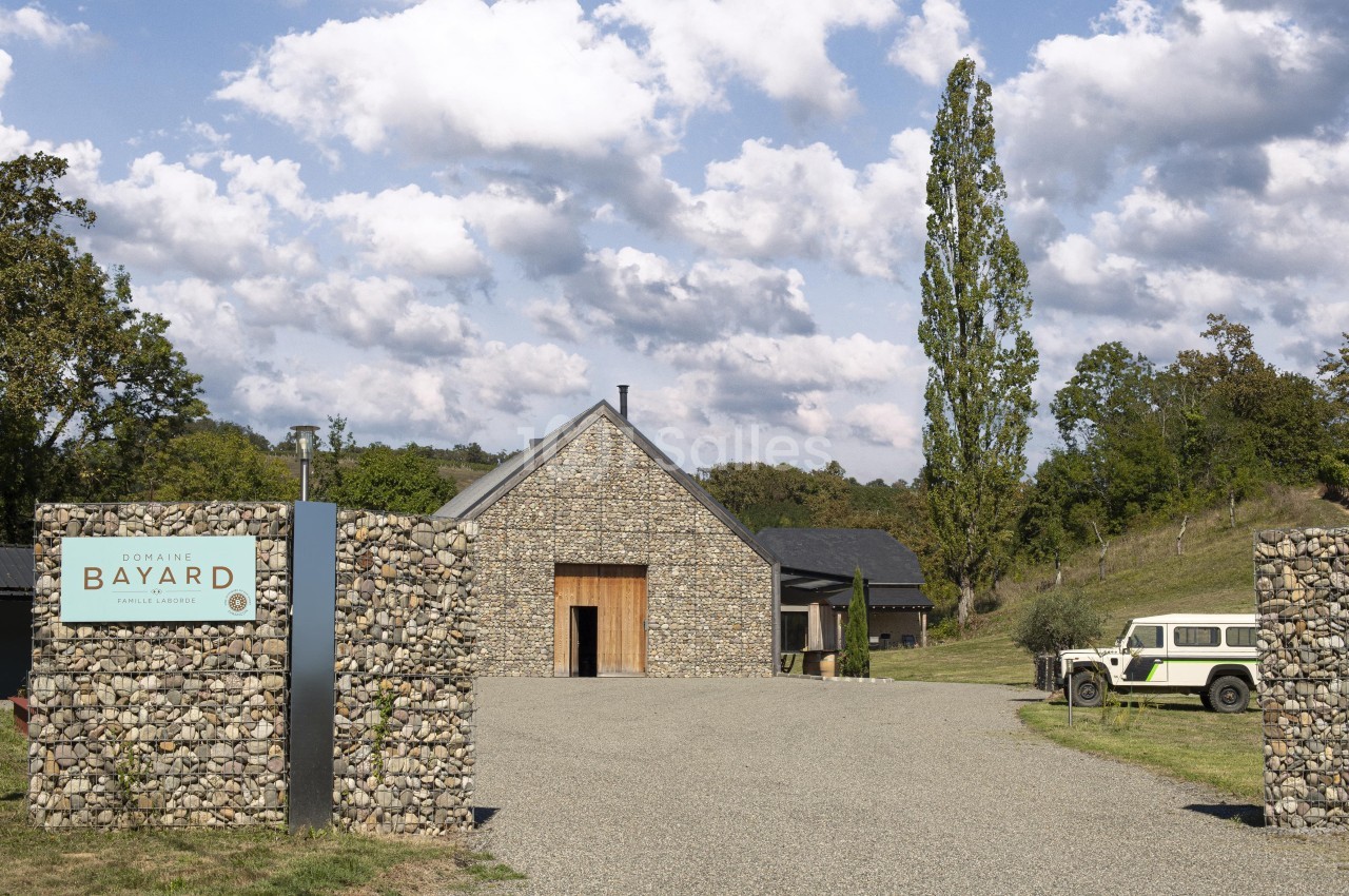 Bâtiment en pierre entouré de verdure avec une entrée gravillonnée et un panneau indiquant ’Domaine Bayard’.