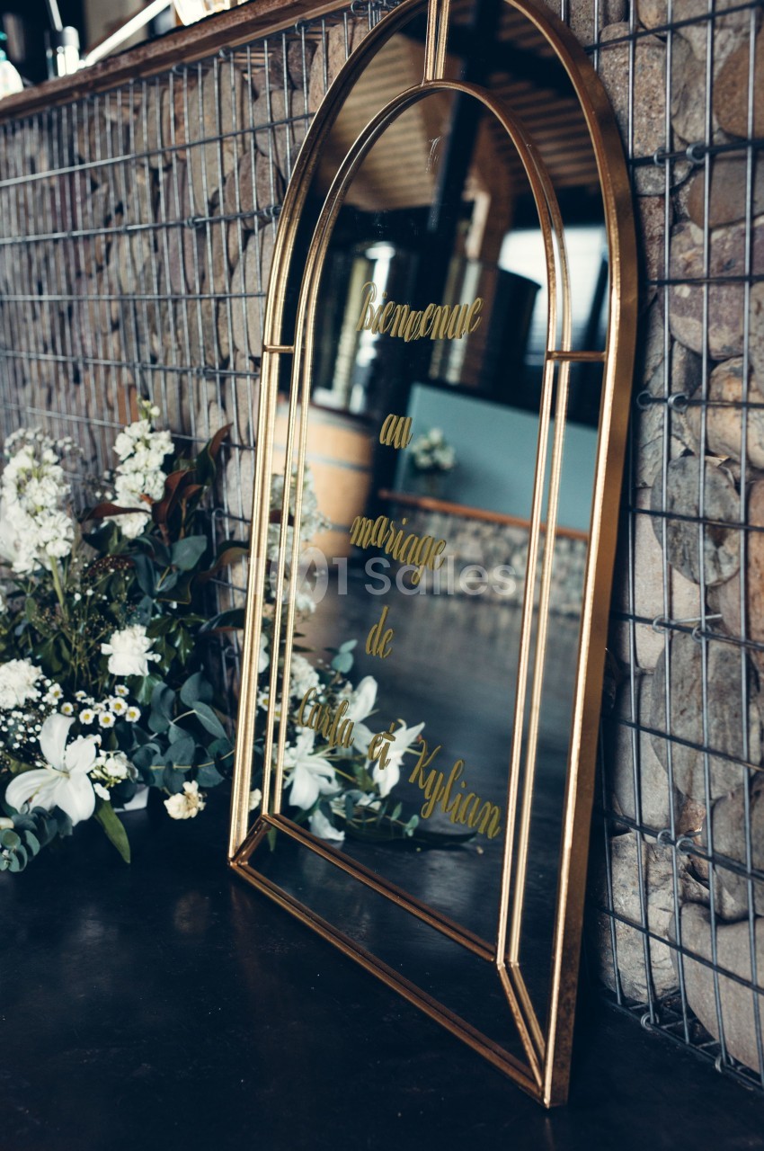 Miroir doré avec inscription ’Bienvenue au mariage de Julia et Kylian’, entouré de fleurs blanches et feuillage.